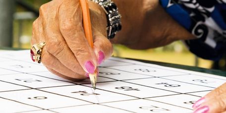 Ältere Frau mit rosa Fingernägeln füllt Bingo-Papier mit einem Bleistift aus