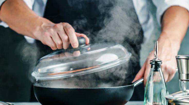 Koch öffnet den Deckel einer dampfenden Pfanne